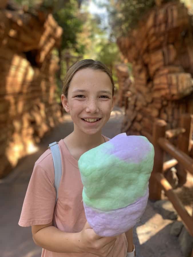 Miss E smiling, surrounded by two rock walls at California Adventure, holding a purple and green gluten-free Halloween cotton candy