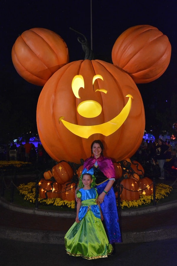 Miss E in a Drizzella costume and Heather in a Princess Anna costume, in front of a winking, Mickey Mouse jack-o-lantern