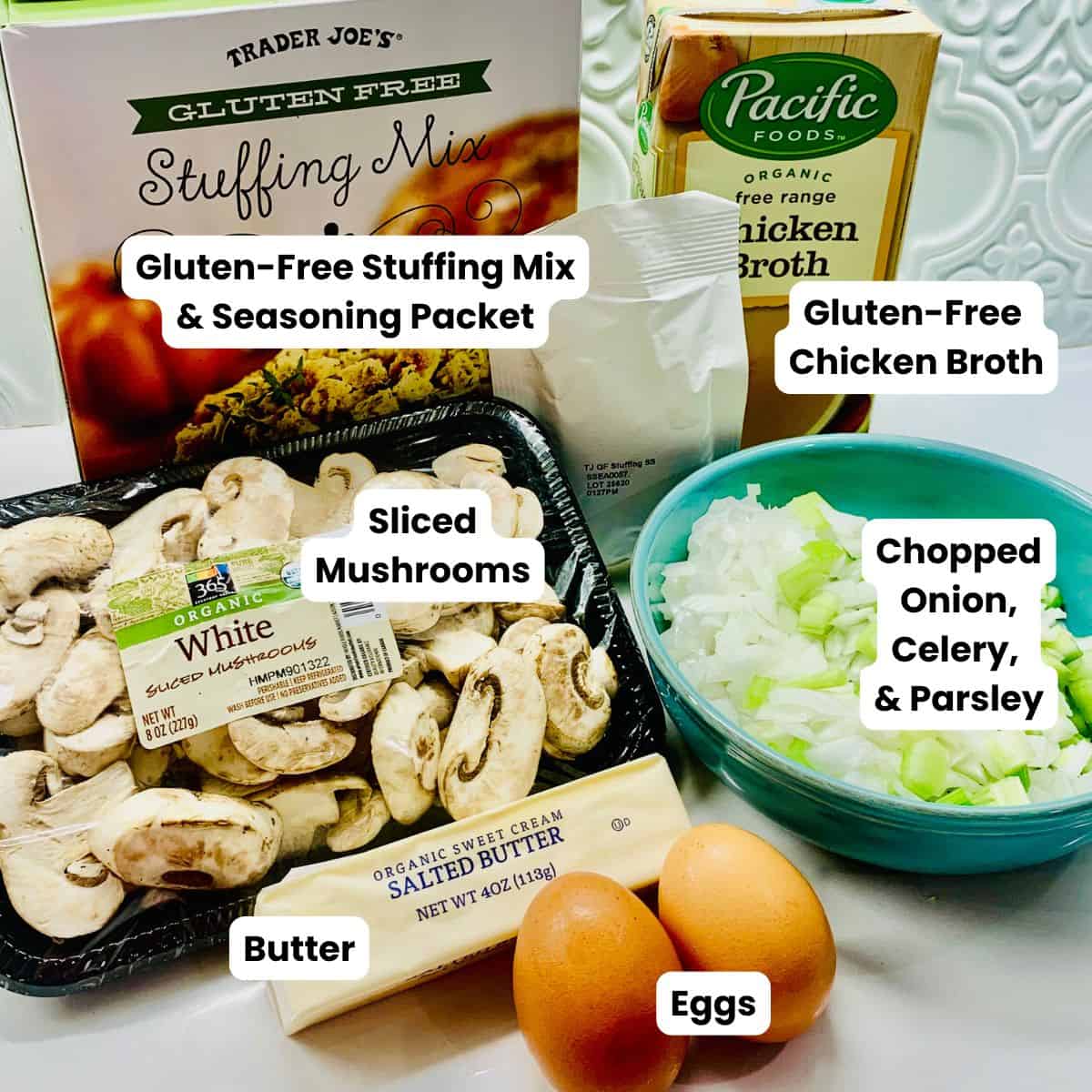 Labeled items with white text box and black text: a box of Trader Joe's gluten-free stuffing mix, the white seasoning packet from the stuffing mix, a box of chicken broth, a package of sliced mushrooms, a blue bowl of chopped onions and celery, a wrapped stick of butter and two brown eggs.