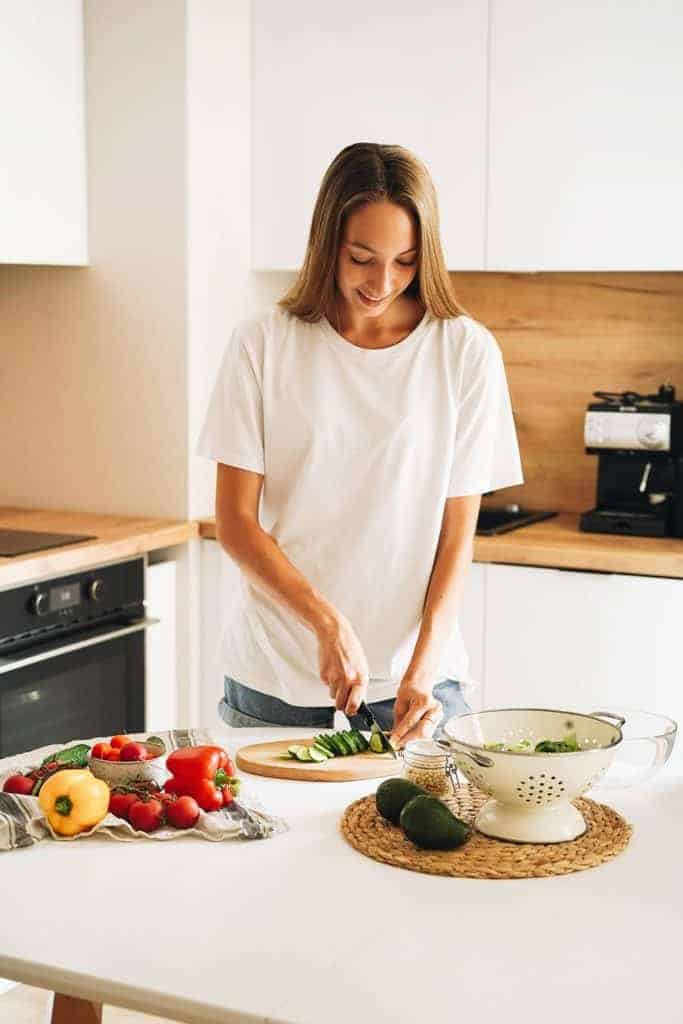 woman in white shirt, in her kitchen chopping cucumber, a white colander with avocados in the foreground and multi-colored bell peppers to her side, all on a white table