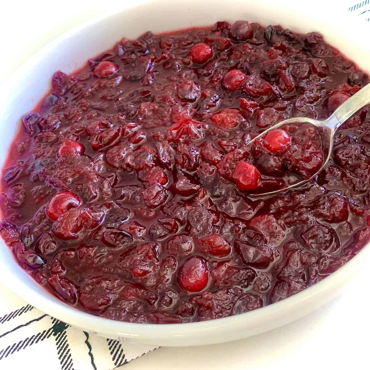 Spoon scooping cranberry sauce out of a white bowl on top of a black and white plaid dishtowel.