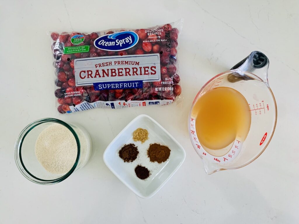 Birds eye view: bag of Ocean Spray Cranberries, glass of white sugar, square white bowl of four spices, and measuring cup with apple cider.