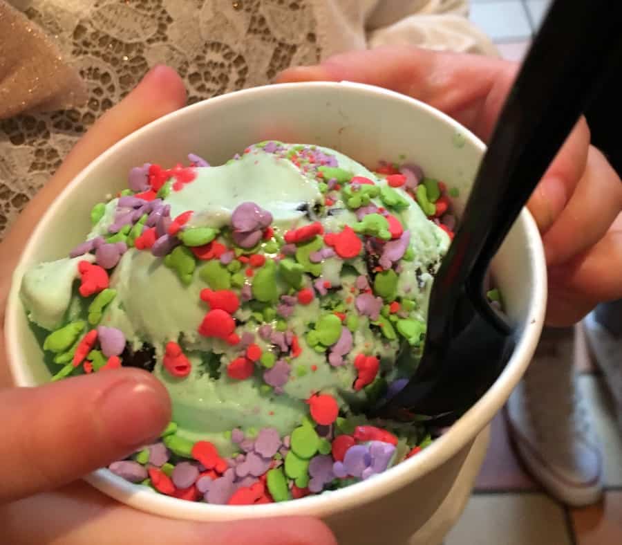 gluten-free mint chip ice cream with Mickey Mouse shaped pink, purple and green sprinkles, with girl's hands holding the cup, and a spoon also in the cup