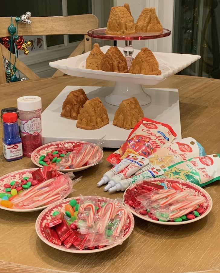 three tiers of gluten-free gingerbread house cakes with candy canes, sprinkles, icing and gluten-free candy on a table in front of the cake tiers