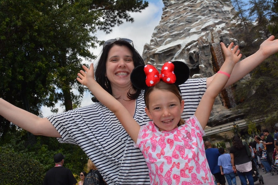 Heather in a stiped navy and white shirt and Miss E in a white shirt with pink butterflies (with Minnie Mouse ears) both raising their arms and smiling in excitement. The Matterhorn is in the background
