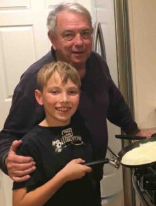 CJ & grandfather smiling together while CJ holds a crepe pan with a gluten-free crepe
