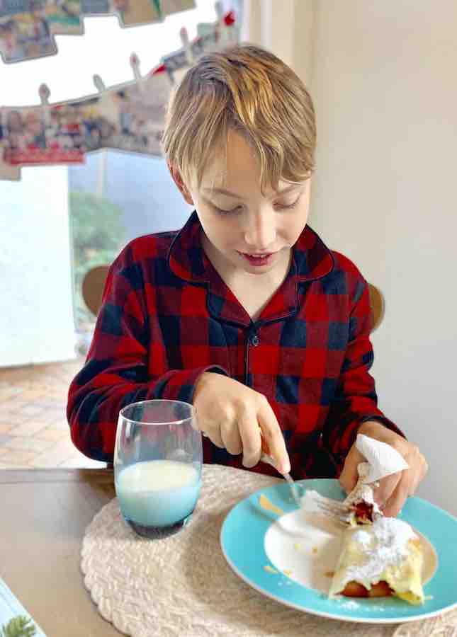 CJ, in black and red checkered PJs with Christmas cards hanging in the background, cutting into his crepe with a fork. A full glass of milk is on the table