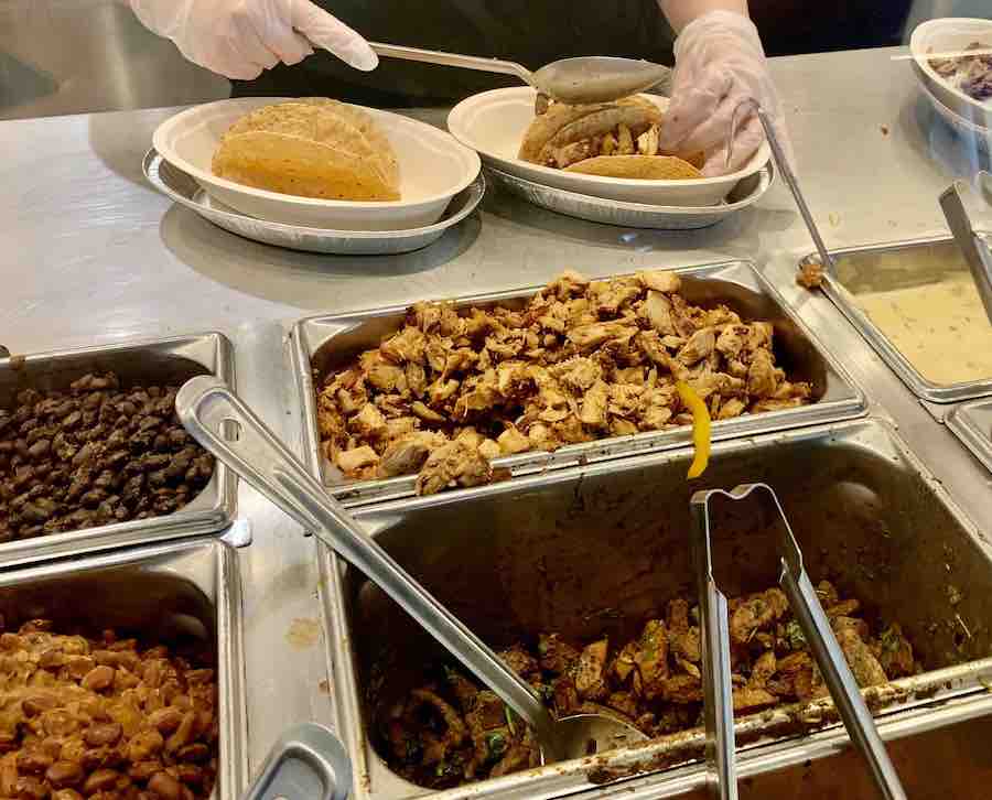 gluten-free meat options at Chipotle in the foreground, employee scooping meat into crispy taco shells in the background
