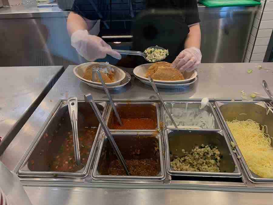 gluten-free salsa options at Chipotle in the foreground, employee scooping salsa onto crispy tacos in the background