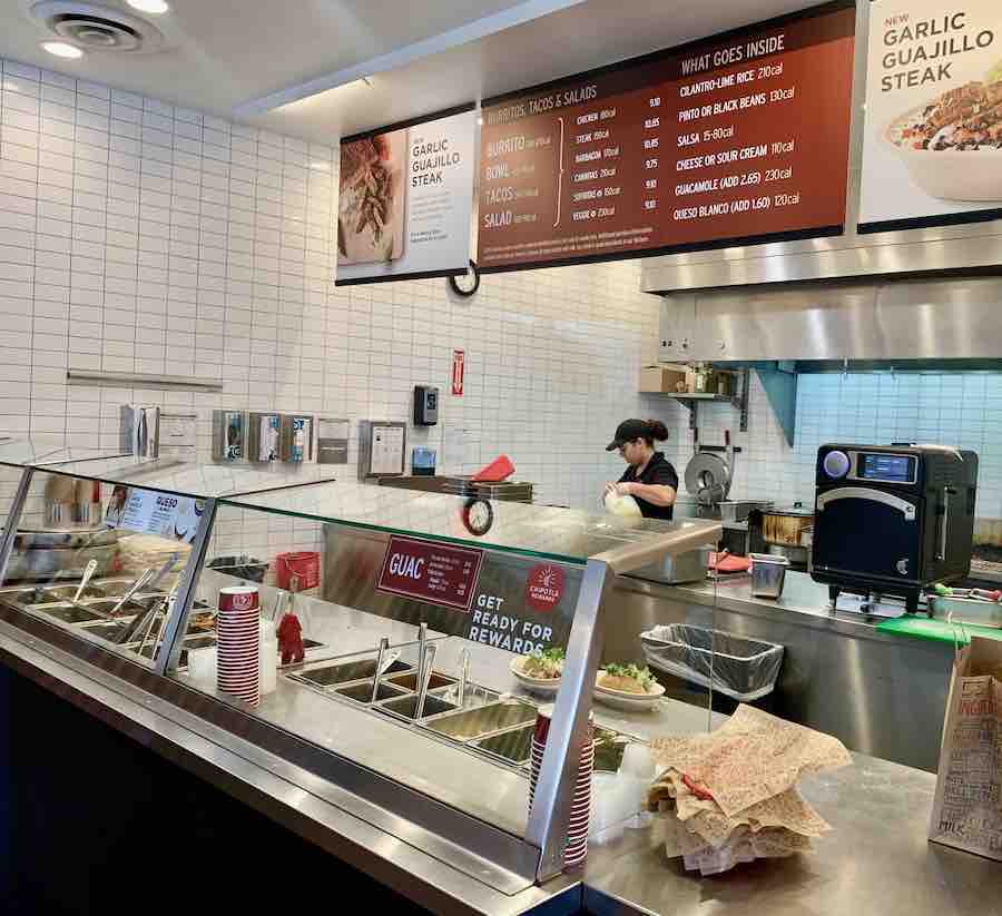 assembly line showing all the gluten-free food at Chipotle, food in the foreground covered by glass, menu above and kitchen in the background
