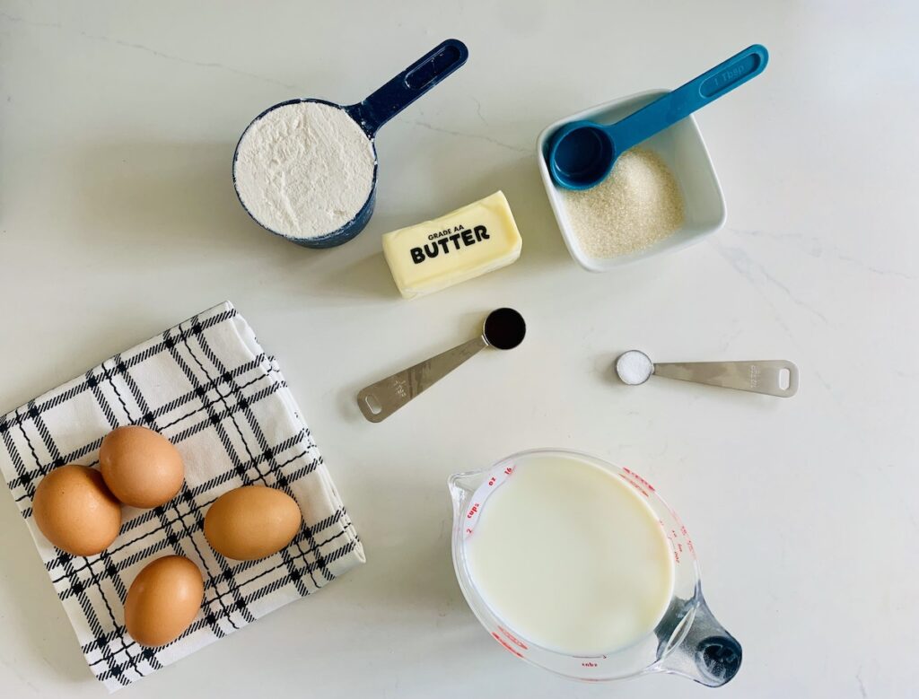 Bird's eye view of ingredients: four brown eggs on a black and white plaid dish towel, a cup of gluten-free flour, a package of butter, a small bowl with measured out sugar and a teal measuring spoon resting in the bowl, two small metal measuring spoons with vanilla and salt, and a liquid measuring cup with milk.