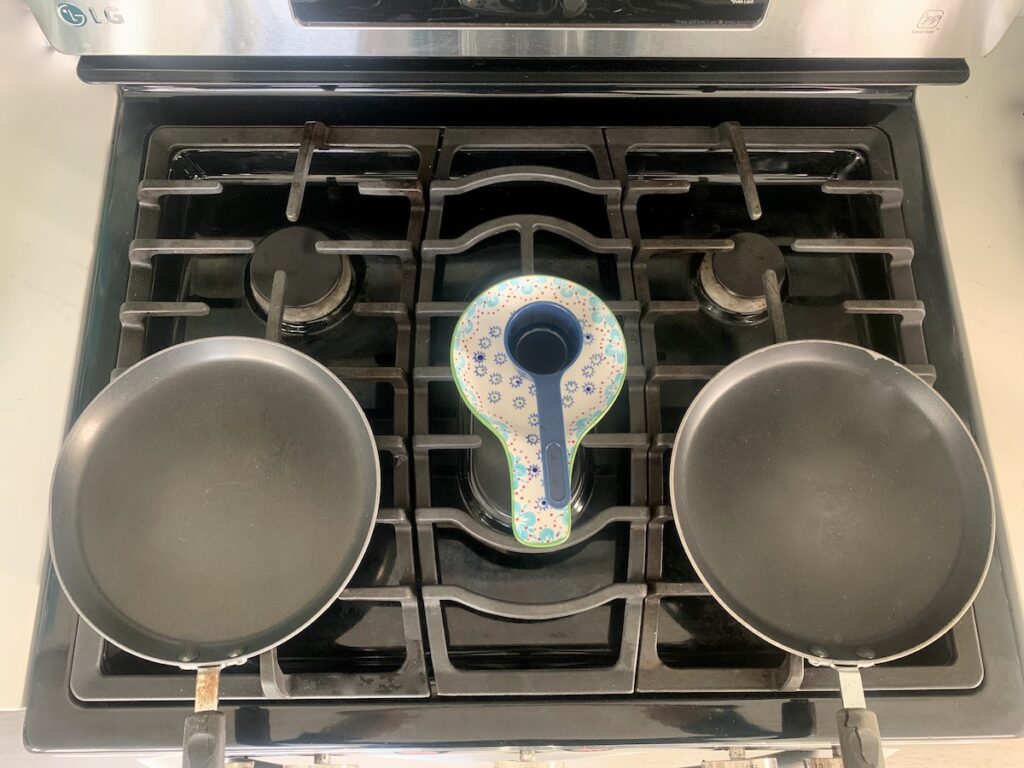 Bird's eye view: 2 crepe pans sitting on a black stove top with a spoon rest between them holding a blue measuring cup.