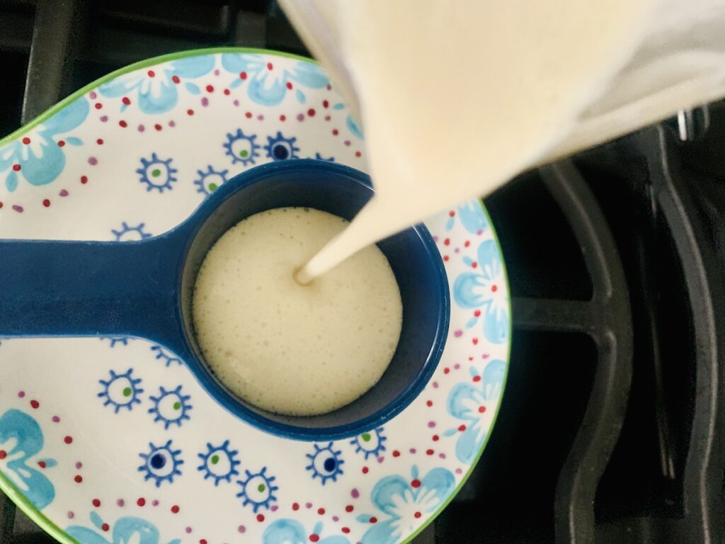 Bird's eye view: crepe batter being poured from the blender into a dark blue measuring cup sitting on a ceramic floral spoon rest. The better looks like thick cream.