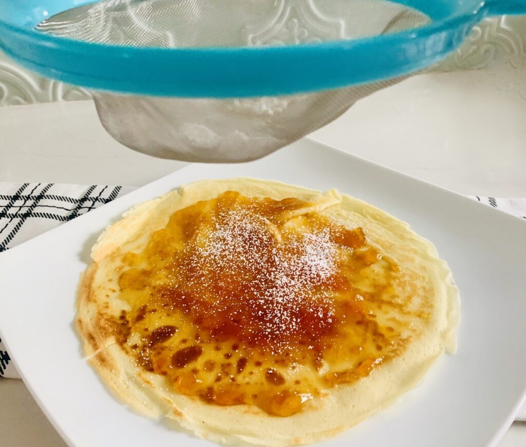 A gluten-free crepe covered in apricot jelly. A flour sifter is above the crepes sprinkling powdered sugar on the crepes.
