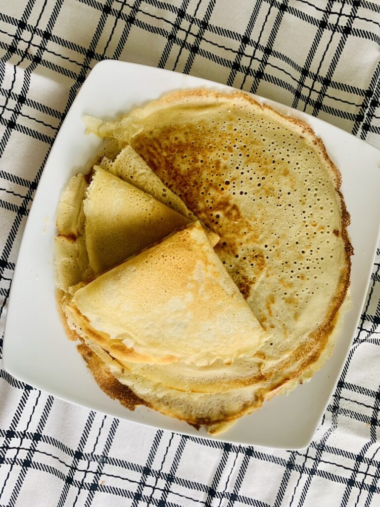 Bird's eye view: a pile of crepes on a square, white plate on top of a black and white plaid dish towel. The top two crepes are folded into triangles.