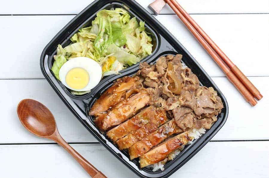 to go container with half a hard-boiled egg and salad in one side, and meat, sauce and rice in the other side, spoon to the left and chopsticks on the right of the container