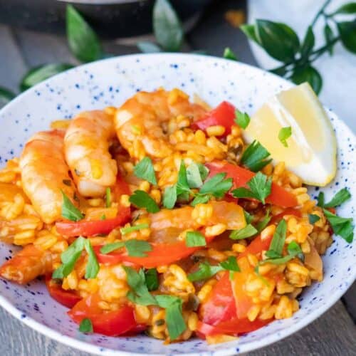 Paella in a white and blue bowl with visible shrimp, orange rice, vegetables, herbs, and lemon slice.