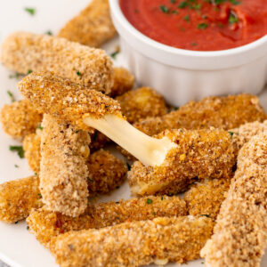 gluten-free mozzarella sticks, one is stretched open revealing a gooey inside, white bowl with marinara dipping sauce