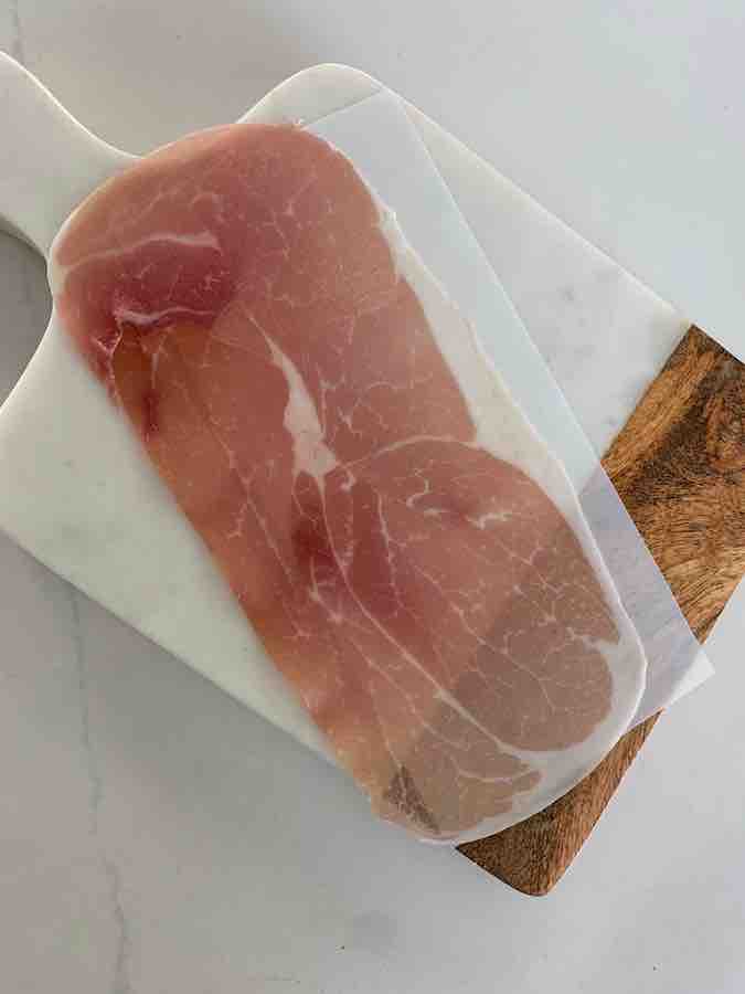 a slice of prosciutto sitting on it's paper divider, on a wood & marble cutting board