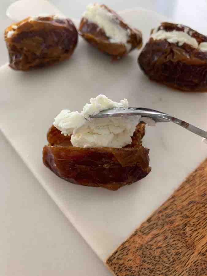 small spoon filling a date with goat cheese, three stuffed dates in the background, all are sitting on a wood & marble cutting board