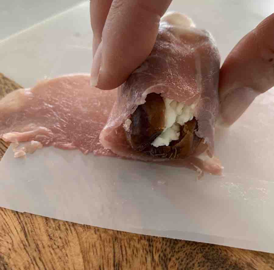 a date stuffed with goat cheese being rolled in a prosciutto strip on it's separating paper, all sitting on a wood & marble cutting board