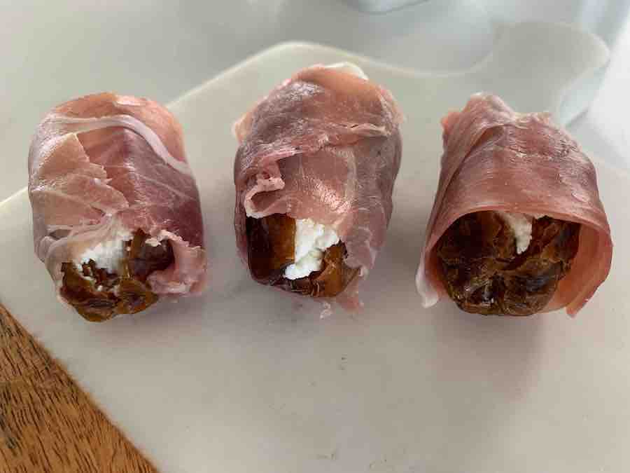three un-baked prosciutto-wrapped dates stuffed with goat cheese, sitting on a wood & marble cutting board