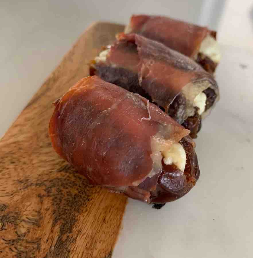row of three baked prosciutto-wrapped dates stuffed with goat cheese, sitting on a wood & marble cutting board