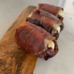 three baked prosciutto-wrapped dates stuffed with goat cheese, sitting on a wood & marble cutting board
