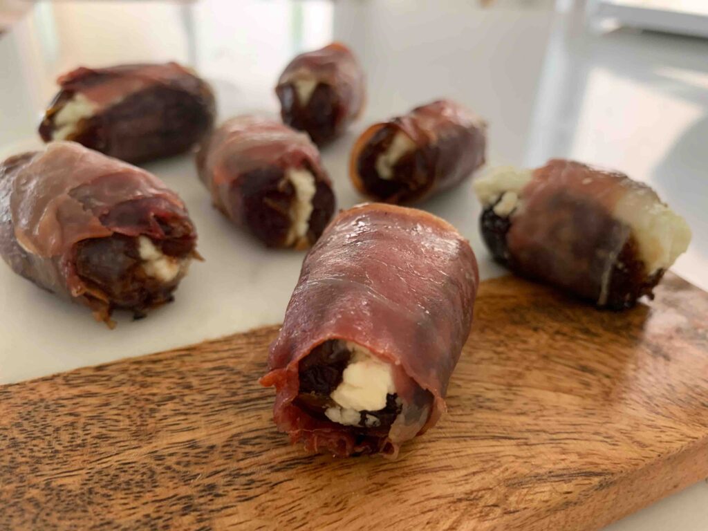 baked prosciutto-wrapped dates stuffed with goat cheese, sitting on a wood & marble cutting board