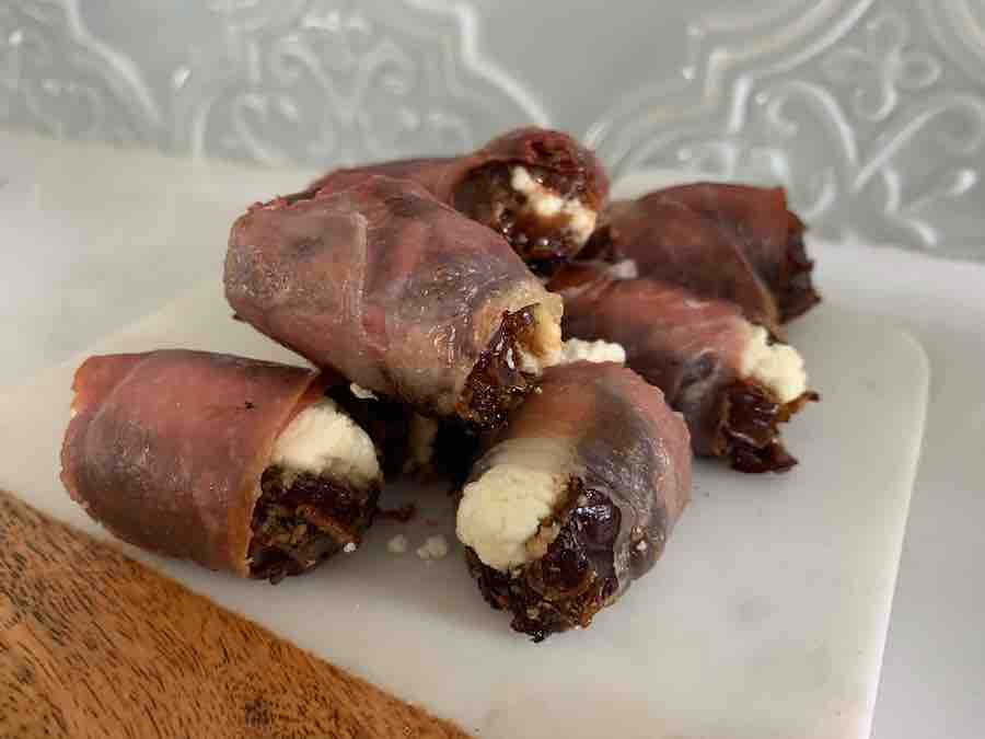 a pile of baked prosciutto-wrapped dates stuffed with goat cheese, sitting on a wood & marble cutting board