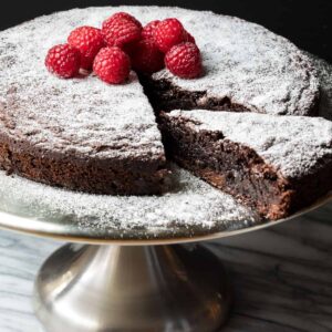 gluten-free chocolate cake with a slice cut out but still on the cake stand, topped with powdered sugar and raspberries