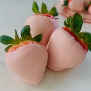 3 pink chocolate covered strawberries with a plate full of more pink chocolate covered strawberries In the background.