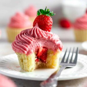 strawberry cupcake with strawberry filling, pink frosting and strawberry on top, inside is showing, sitting on a plate with a fork on plate as if it was cut into.... more cupcakes blurred in background
