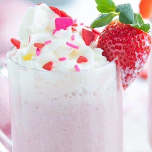 glass mugs with pink hot chocolate, whipped cream, Valentine's sprinkles and strawberry for garnish