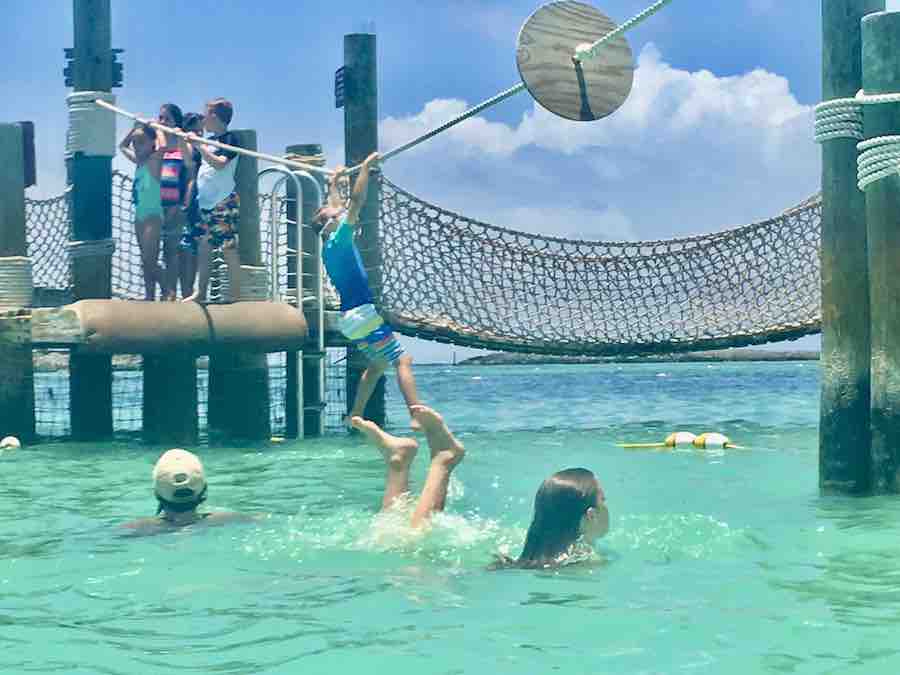 CJ crossing rope by hand, hanging over the ocean, rope bridge and dock for kids in the background, kids playing in the ocean in the foreground, one kid upside down in water with feet sticking up