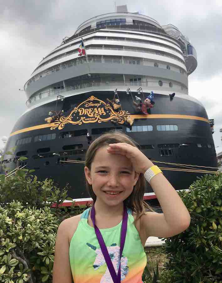 Miss E with her hand blocking the sun, smiling with the Disney Dream cruise ship in the background