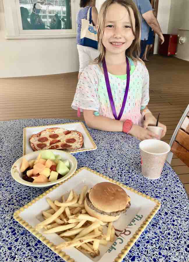 Miss E smiling behind a table full of gluten-free pepperoni pizza, bowl of sliced fruit, gluten-free cheeseburger & fries
