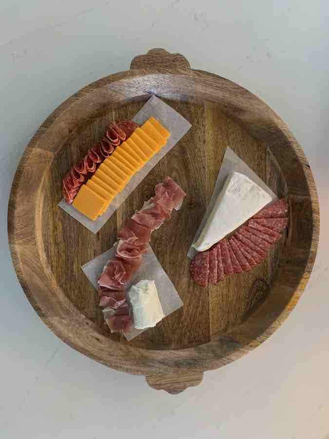 a round, wooden tray with a partially assembled gluten-free charcuterie display with cheese and fold meats