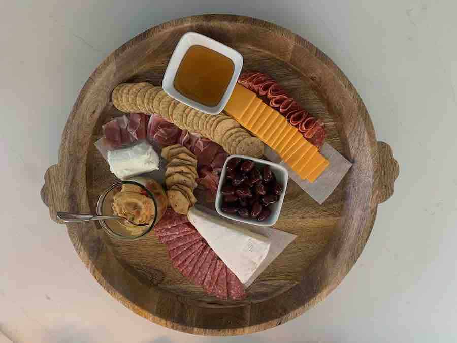 a round, wooden tray with a partially assembled gluten-free charcuterie display: crackers, sliced cheddar, honey, olives, hummus, brie and goat cheese