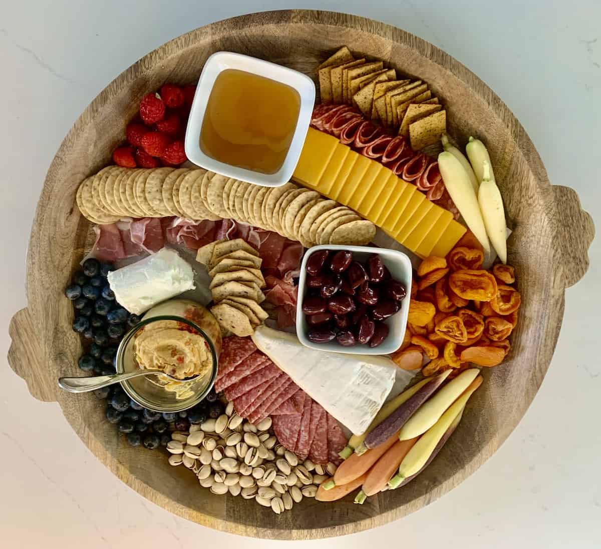 top view of of a round gluten-free charcuterie board with crackers, apricots, sliced cheddar salami, prosciutto, honey, olives, hummus, pistachios, raspberries, blueberries, brie and goat cheese
