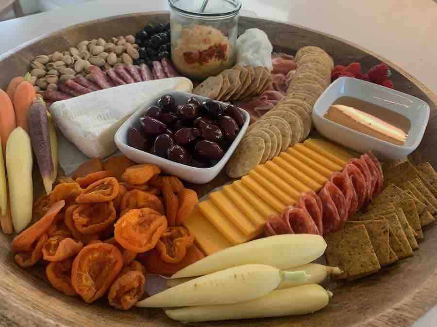 side view of of a round gluten-free charcuterie board with crackers, apricots, sliced cheddar salami, prosciutto, honey, olives, hummus, pistachios, raspberries, blueberries, brie and goat cheese