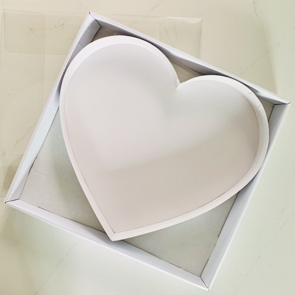 Square white box with heart shaped box inside. Plastic top lying flat partially under the box.