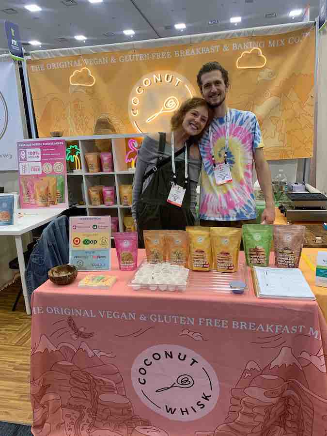 two trade show reps smiling (one leaning into the other behind a table of baking mixes... Pink tablecloth with logo for Coconut Whisk company