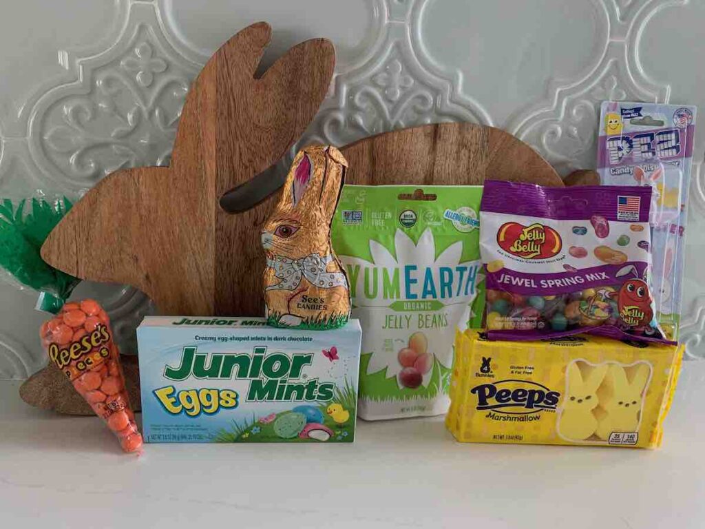 a wooden easter bunny backdrop with Easter candy in front: Reese's pieces in carrot packaging (positioned as if the bunny were eating the carrot), Junior Mints Eggs, See's Hollow Chocolate Bunny, Yum Earth Jelly Beans, Jelly Belly Jelly Beans, Peeps Bunnies, Easter Bunny PEZ dispenser