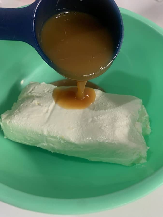 caramel syrup being poured from a measuring cup onto softened cream cheese