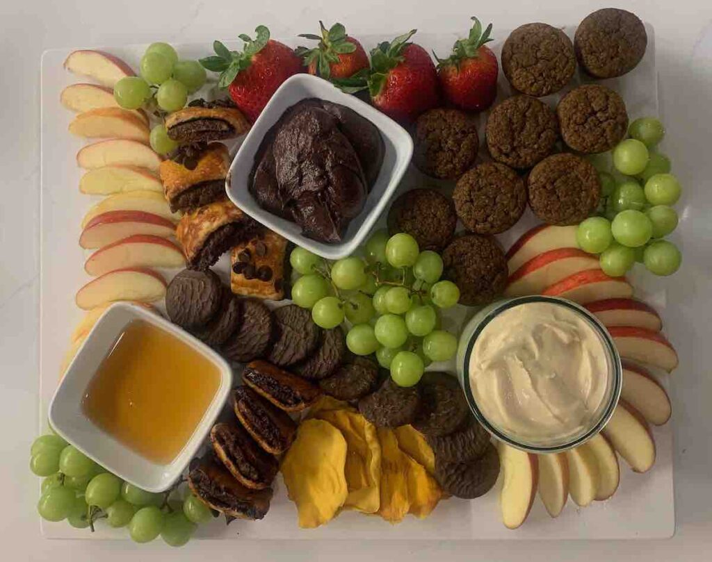 gluten-free dessert charcuterie with: cream cheese caramel dip, honey, chocolate hummus, red apple slices, green grapes, dried mango, banana muffins, chocolate rugelach, and strawberries