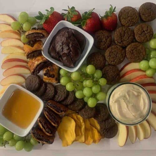 gluten-free dessert charcuterie with: cream cheese caramel dip, honey, chocolate hummus, red apple slices, green grapes, dried mango, banana muffins, chocolate rugelach, and strawberries