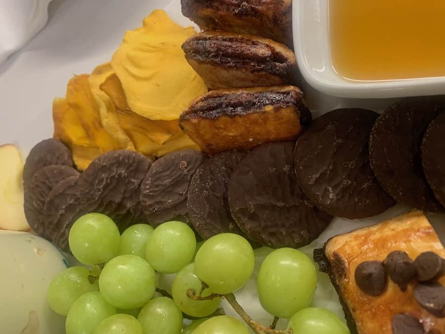 close up of part of a gluten-free dessert charcuterie, including: Goodie Girl Mint Slims, dried mango, chocolate mango, green grapes and a corner of a bowl of honey