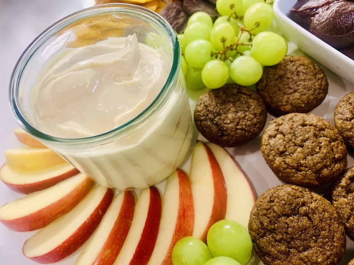 Caramel Cream cheese dip in a glass surrounded by sliced apples, green grapes and mini muffins