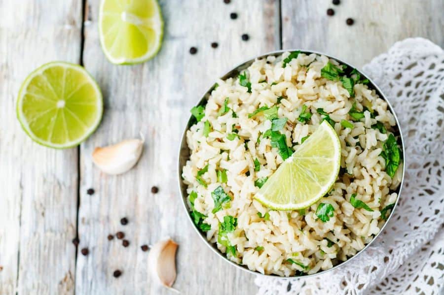 cilantro-lime rice in a bowl on a table, also on the table are cut limes, garlic, black peppercorns and white lace
